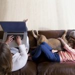 photo of mom and daughter on couch with mom on laptop and girl laying down with dog