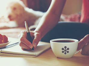 Picture of woman writing in a notebook holding a cup of coffee. Crossiants are on a plate on the table.