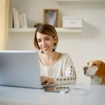 Picture of woman sitting in front of laptop with dog sitting next to her on a chair